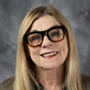 Smiling woman with long blonde hair and black thick-framed glasses, posed against a gray studio backdrop.